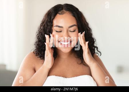 Eine glückliche, übergroße Frau, die ihr Gesicht berührt und mit geschlossenen Augen lächelt und zu Hause Schönheitsbehandlungen macht Stockfoto