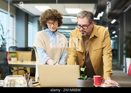 Ein erwachsener Mann und eine junge Geschäftsfrau sehen sich ein Online-Webinar oder eine Konferenz an, während sie sich über das Notebook beugen und auf den Bildschirm schauen Stockfoto