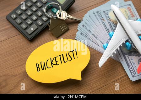 Draufsicht auf Flugzeugmodell, Geld, Hausschlüssel, Taschenrechner und gelbe Sprache geschrieben mit Bucketlist auf Holzhintergrund mit Kopierraum. Stockfoto