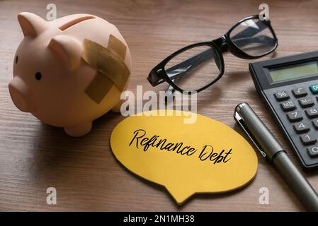 Selektiver Fokus auf Sparschwein, Brille. Taschenrechner, Stift und gelbe Sprechblase mit Refinanzierungsschulden auf Holzhintergrund. Stockfoto