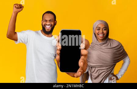 Ein überfreuliches schwarzes muslimisches Paar, das ein großes, leeres Smartphone mit leerem Bildschirm zeigt Stockfoto