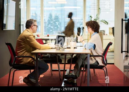 Seitenansicht von zwei Kollegen, die am Tisch sitzen und Laptop und Smartphone gegen die unscharfe Kontur eines vorbeigehenden Mannes verwenden Stockfoto