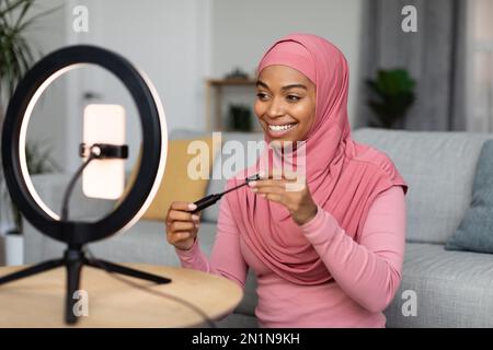 Schönheitsblogger. Glückliche afroamerikanische Dame in Hidschab, die neue Mascara sieht, auf ein Smartphone mit Ringlampe schaut Stockfoto