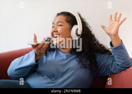 Glückliche hispanische Dame, die auf dem Sofa sitzt, Lieder singt, telefoniert Stockfoto