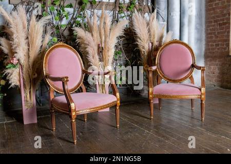 Inneneinrichtung des Wohnzimmers mit pinkem Sessel auf Holzboden. Stilvolle pinke Sessel und Pampasgras in einer Vase, helles minimalistisches Interieur Stockfoto