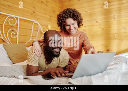 Eine junge lächelnde Frau umarmt ihren Mann und zeigt auf den Laptop-Bildschirm, während sie sich im Bett entspannt und Online-Videos ansieht Stockfoto