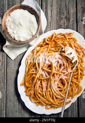 Bucatini: Rezept mit Amatriciana-Sauce, mit Parmesan. Grauer rustikaler Holztisch, weiße Platte, Schüssel mit Parmesan und weißes Geschirrtuch. Vertikale Beschriftung. Stockfoto