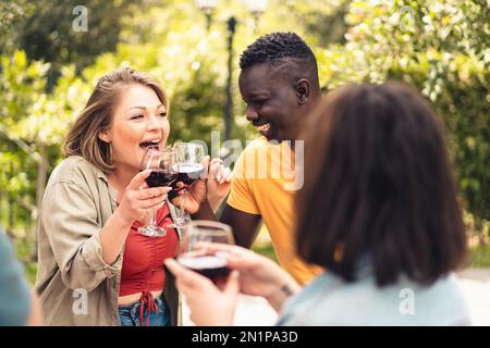 Eine kurvige junge Erwachsene Kaukasierin und ein junger afrikanischer Mann jubeln und lachen, während sie mit Rotwein anstoßen - vielfältige fröhliche Menschen Lebensstil Conc Stockfoto