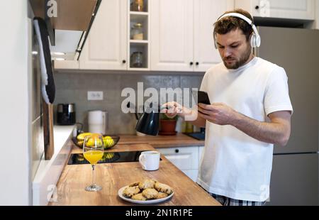 Mann liest Nachrichten im Internet mit seinem Handy, während er morgens in der Küche Kaffee kocht. Stockfoto
