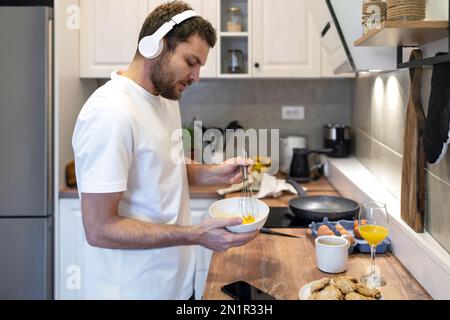 Mann mit weißem T-Shirt und Kopfhörern steht in der Küche und kocht Omelett zum Frühstück und hört Musik. Stockfoto
