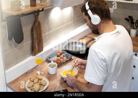 Mann mit weißem T-Shirt und Kopfhörern steht in der Küche und kocht Omelett zum Frühstück und hört Musik. Stockfoto