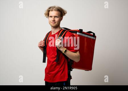 Lächelnder Kurierfahrer mit lockigem Haar in roter Uniform und isoliertem Thermobeutel auf weißem Hintergrund. Lieferservice. Stockfoto