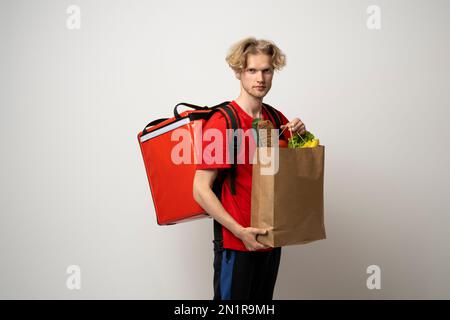 Ein attraktiver Zusteller, ein Kurier, der Lebensmittel und Getränke aus dem Laden mitbrachte, ein Supermarkt, isoliert auf weißem Studiohintergrund. Stockfoto