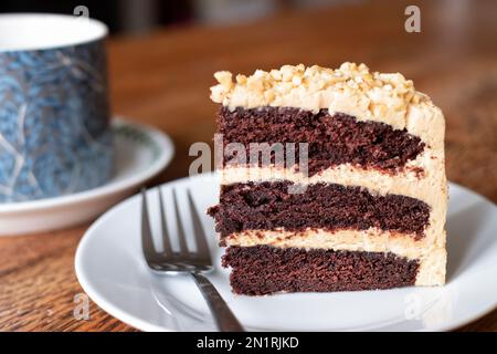 Ein Stück frisch gebackene Erdnussbutter und Schokoladenkuchen. Der Kuchen ist mit einer dicken Buttercreme gefüllt und wird zusammen mit einer Tasse Tee auf einem Beilagenteller serviert Stockfoto