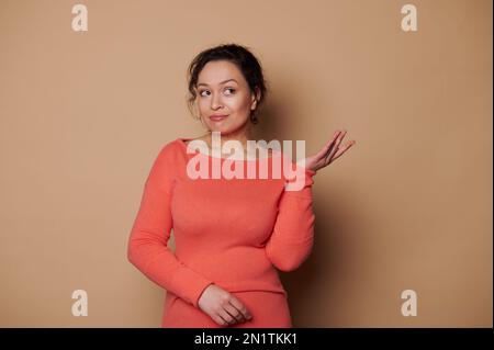 Eine nachdenkliche Frau in leuchtend orangefarbenem Kleid, die Überraschung ausdrückt und auf Werbeschriften auf beigefarbenem Hintergrund zeigt Stockfoto