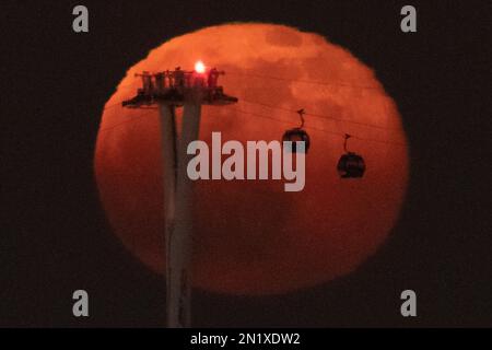 London, Großbritannien. 6. Februar 2023 UK Weather: 99,1 % Vollmondaufgang erzeugt Silhouetten von vorbeifahrenden Seilbahnen, auch als IFS Cloud Cable Car im Osten Londons bekannt. Kredit: Guy Corbishley/Alamy Live News Stockfoto