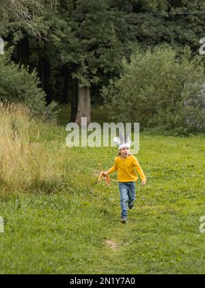 Der Laufbursche spielt auf dem Spielfeld Indianer. Der Junge hat einen handgemachten Kopfschmuck aus Federn und eine Schleife mit Pfeilen. Kostümrollen. Outdoor Freizeit A Stockfoto