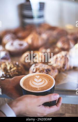 Frauenhände halten eine Tasse Kaffee, zubereitet von einem Barista, neben einer Theke voller Croissants. Restaurant. Ruhige Atmosphäre. Frühstück. W Stockfoto