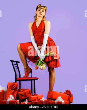 Frau in rotem Kleid mit Retro-Frisur und einem Strauß Tulpen. Hübsches Mädchen mit Blumenstrauß und Geschenkbox. Valentinstag, Geburtstag, Frauen Stockfoto