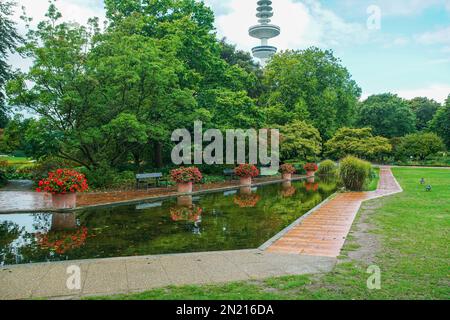 Ein Teich mit roten Blumen in großen Töpfen, der sich im Wasser im botanischen Garten 'Planten un Blomen' in Hamburg widerspiegelt. Fernsehturm in Hamburg, gar Stockfoto