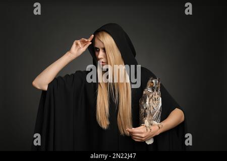 Hexe in schwarzem Mantel mit Eule auf dunklem Hintergrund. Unheimliche Fantasie-Figur Stockfoto