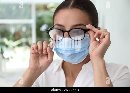 Frau, die neblige Brille abwischte, verursacht durch das Tragen einer medizinischen Maske in Innenräumen, Nahaufnahme Stockfoto