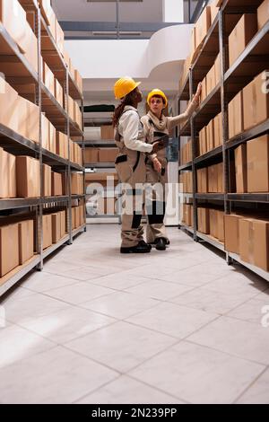 Mitarbeiter des Versandzentrums scannen Kartons vor dem Transport. Zwei Lagerarbeiterinnen tragen Schutzhelme und Overalls, die sich unterhalten, stehen neben Regalen voller Kartons Stockfoto