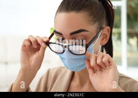 Frau, die neblige Brille abwischte, verursacht durch das Tragen einer medizinischen Maske in Innenräumen, Nahaufnahme Stockfoto