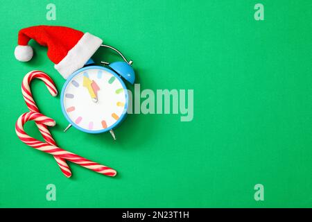 Wecker mit Weihnachtsmannmütze und Zuckerstangen auf grünem Hintergrund, flach liegend mit Platz für Text. Der Countdown für das neue Jahr Stockfoto