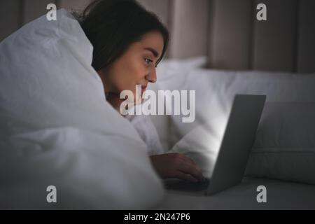 Junge Frau mit Nomophobie, die nachts einen Laptop im Bett benutzt. Insomnie-Konzept Stockfoto