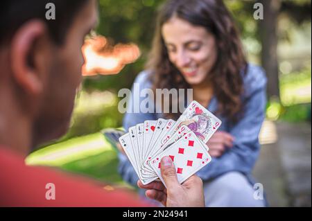 Kartenspiel in einem Frühlingsgarten: Das Paar sitzt auf einem Tisch, entspannt und spielt Karten zusammen. Unscharfer Hintergrund. Stockfoto