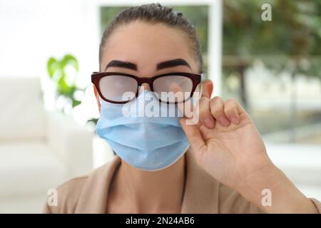 Frau, die neblige Brille abwischte, verursacht durch das Tragen einer medizinischen Maske in Innenräumen, Nahaufnahme Stockfoto