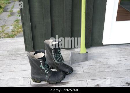 Alte schwarze Stiefel und Axt mit leuchtend grünem Axtgriff an Deck des Schuppens Stockfoto