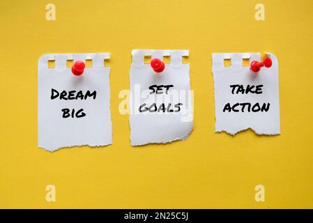 Drei Papierstücke mit Notizen TRÄUMEN VON GROSSEN TRÄUMEN, SETZEN ZIELE UND ERGREIFEN MASSNAHMEN auf gelbem Hintergrund. Stockfoto