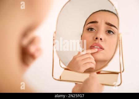 Emotionale Frau mit Herpes berührt Lippen vor dem Spiegel vor hellem Hintergrund Stockfoto