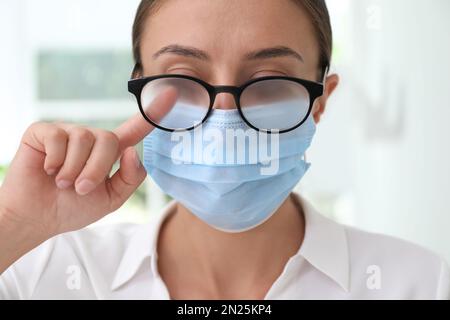 Frau, die neblige Brille abwischte, verursacht durch das Tragen einer medizinischen Maske in Innenräumen, Nahaufnahme Stockfoto