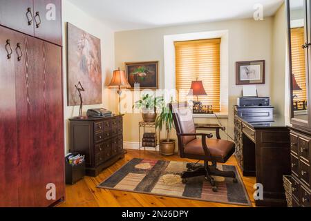 Home Office mit herrschaftlich braunem Ledersessel und Schreibtisch in einer Ecke des Wohnzimmers im alten, renovierten Haus im Canadiana Cottage Stil aus dem Jahre 1840. Stockfoto