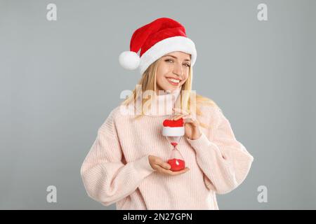 Frau mit Weihnachtsmannmütze mit Sanduhr auf grauem Hintergrund. Der Countdown für Weihnachten Stockfoto