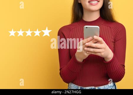 Frau, die ihre Bewertung online über ein Smartphone auf gelbem Hintergrund mit fünf Sternen abgibt, Nahaufnahme. Service- oder Produkt-Feedback Stockfoto