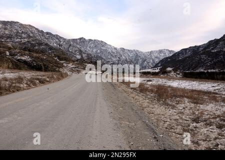 Eine gerade Schotterstraße durch ein schneebedecktes Tal zwischen Bergketten an einem klaren Wintertag. Altai, Sibirien, Russland. Stockfoto