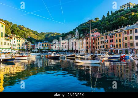 Boote ankern im Hafen von Portofino, hinter ihnen pastellfarbene Fassaden von Portofino, Dampfpfade am blauen Himmel, Portofino, Ligurien Stockfoto