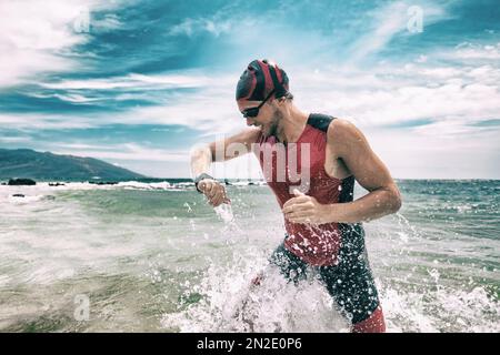 Triathleschwimmer, der während des Triathlons mit Smartwatch auf die Sport Watch App schaut. Schwimmender Mann, der aus dem Meer rennt, schwimmt und die Herzfrequenz auf Smart überprüft Stockfoto