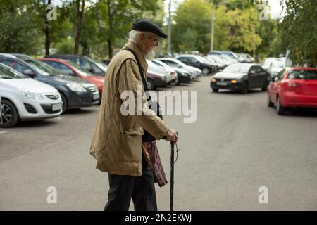 Der alte Mann geht durch den Parkplatz. Alter Mann mit Stock. Mann in armer Kleidung. Pensioniert in Russland. Russischer alter Mann. Stockfoto