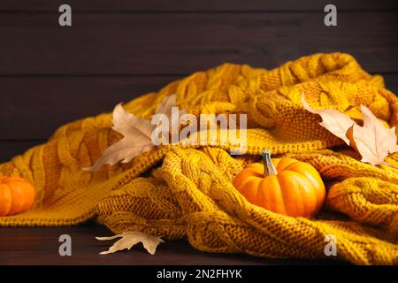 Orangefarbenes Strickmuster mit trockenen Blättern und Kürbissen auf braunem Holztisch, Nahaufnahme Stockfoto