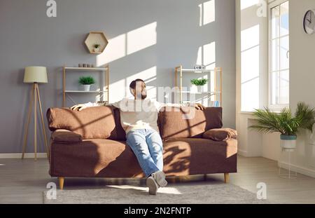Glücklicher junger Mann, der auf einer komfortablen braunen Couch in einem modernen Wohnzimmer sitzt Stockfoto