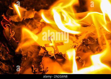 Flammen auf einem brennenden silbernen Kreuz, die in der Nacht glühen. Nahaufnahme brennendes Papier mit silbernem Kreuz in der Mitte auf schwarzem Hintergrund. Stockfoto