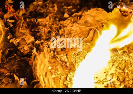 Flammen auf einem brennenden silbernen Kreuz, die in der Nacht glühen. Nahaufnahme brennendes Papier mit silbernem Kreuz in der Mitte auf schwarzem Hintergrund. Stockfoto