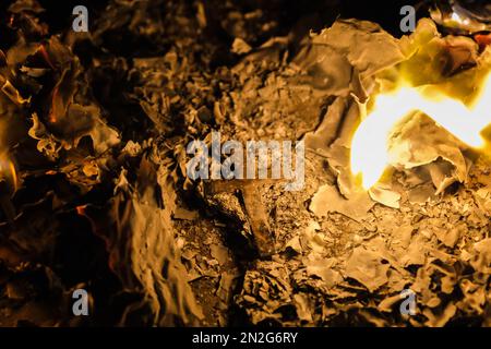 Flammen auf einem brennenden silbernen Kreuz, die in der Nacht glühen. Nahaufnahme brennendes Papier mit silbernem Kreuz in der Mitte auf schwarzem Hintergrund. Stockfoto