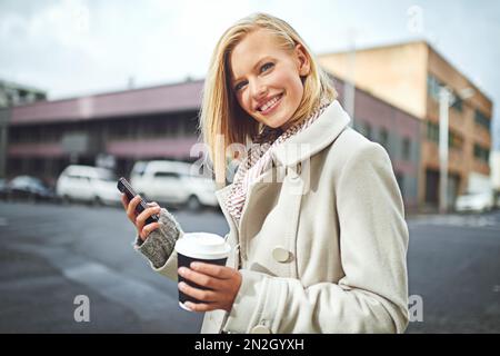 Mir geht es nur um das Stadtleben. Abgeschnittenes Porträt einer attraktiven jungen Frau, die eine SMS schickt, während sie einen Kaffee auf den Straßen der Stadt trinkt. Stockfoto