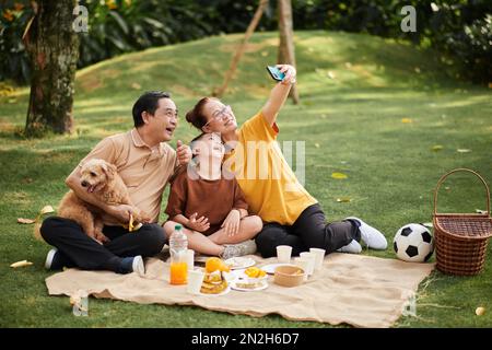 Fröhliche Großeltern machen Selfie mit Hund und Enkel beim Picknick Stockfoto